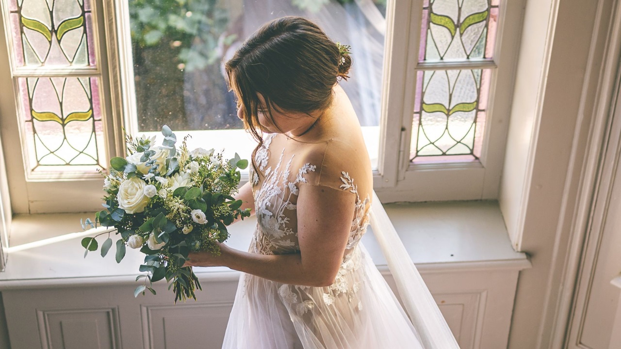 Braut in weissem Kleid mit Brautstrauss steht vor einem Fenster
