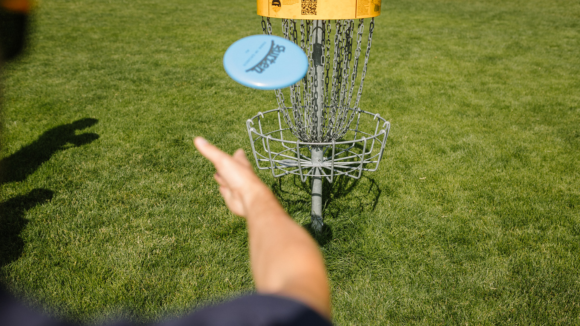 A person throwing a blue disc golf frisbee into the basket