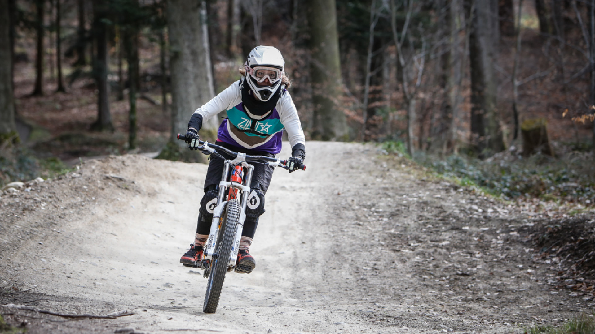 A downhill cyclist riding down the forest trail. The person is wearing bright clothing with a helmet and goggles. 