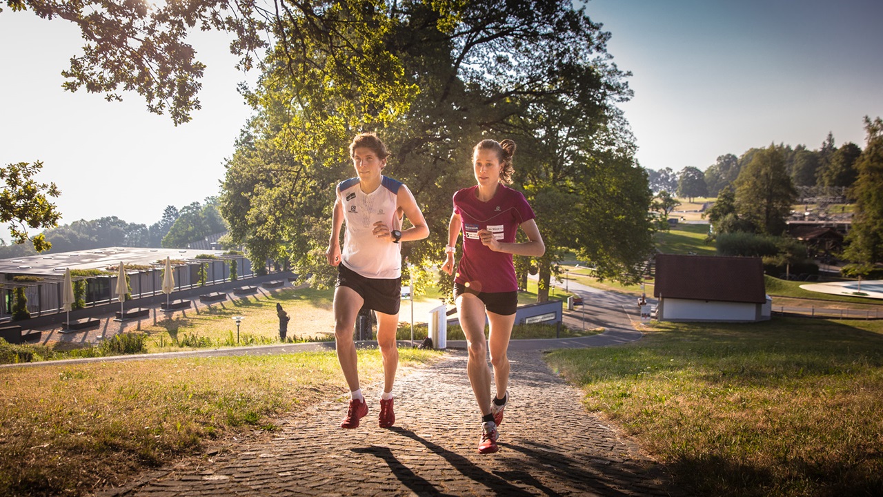 Eine Sportlerin und ein Sportler joggen den Asphaltweg hinauf. 