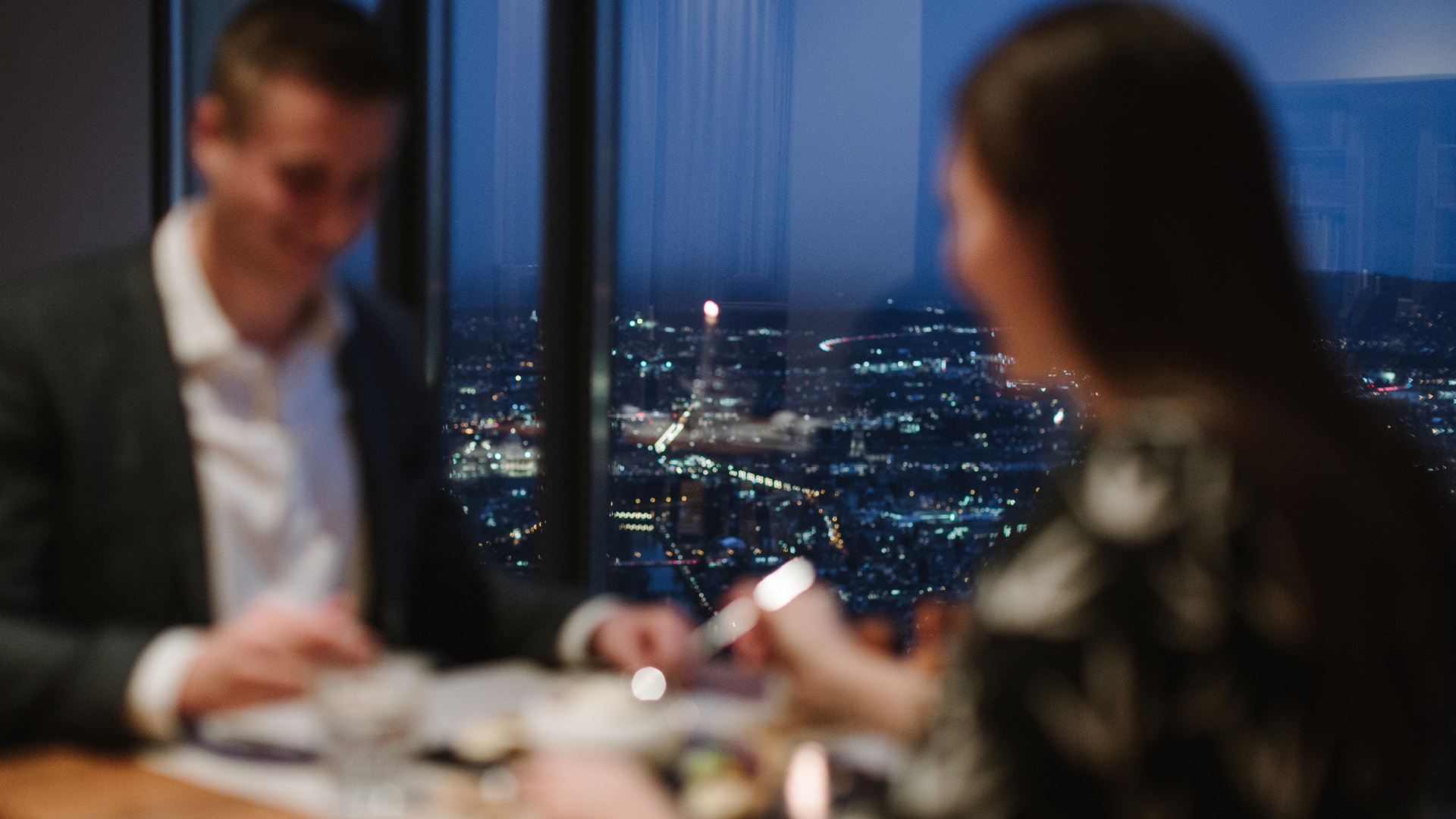 A couple enjoying dinner in a cozy atmosphere. The city lights can be seen in the background. 
