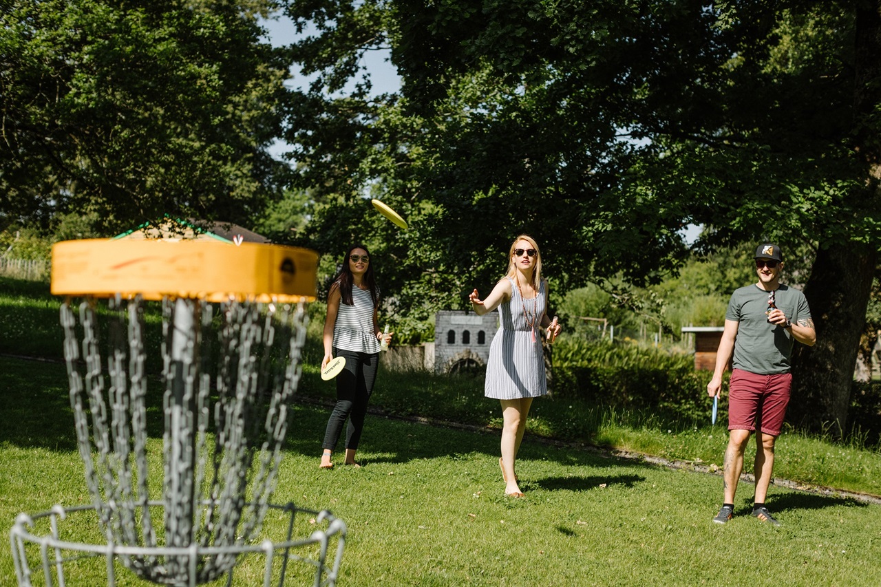 Eine Gruppe spielt Discgolf. Eine Frau wirft die Frisbee-Scheibe ins Ziel. 