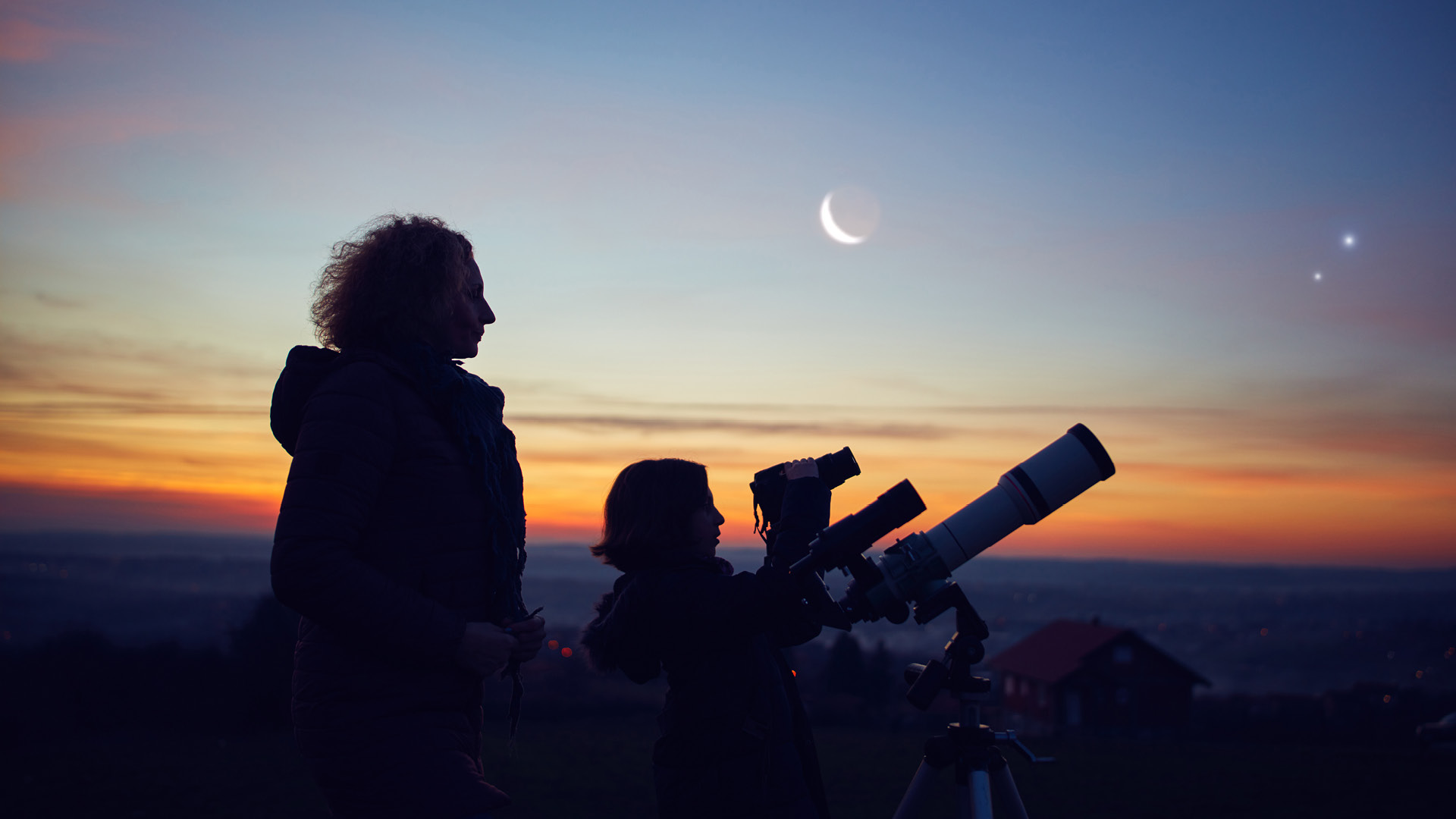 Eine Frau und ein Kind schauen in den Himmel. Ein Teleskop steht vor ihnen. Es ist eine wunderschöne Abendstimmung mit sichtbarem Mond und Sterne. Die Galaxy ist zum Greifen nah. 
