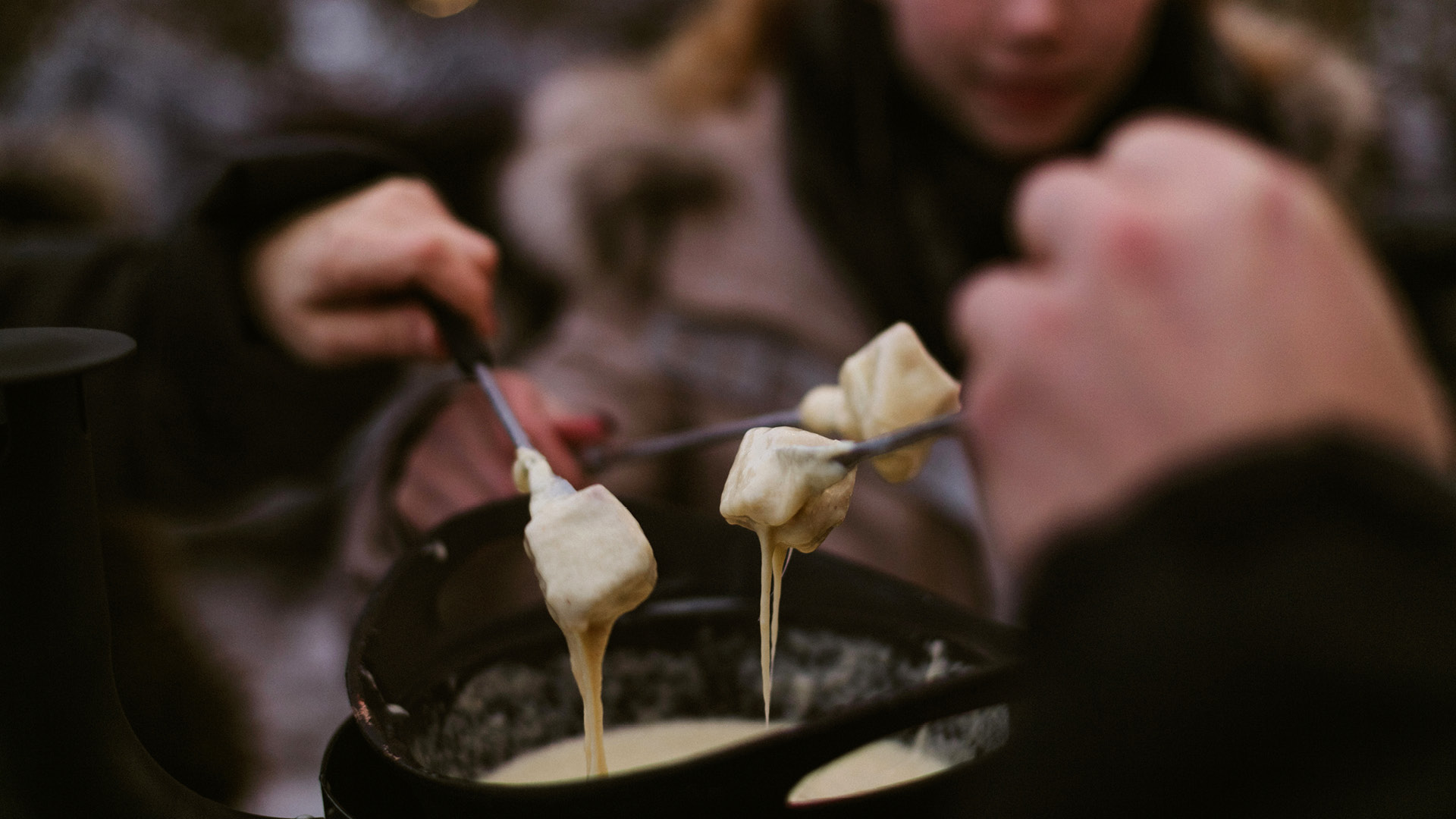 The melted strings of cheese are pulled in a fondue pot. Four forks turn the bread in the pan. There is a wintery atmosphere. 