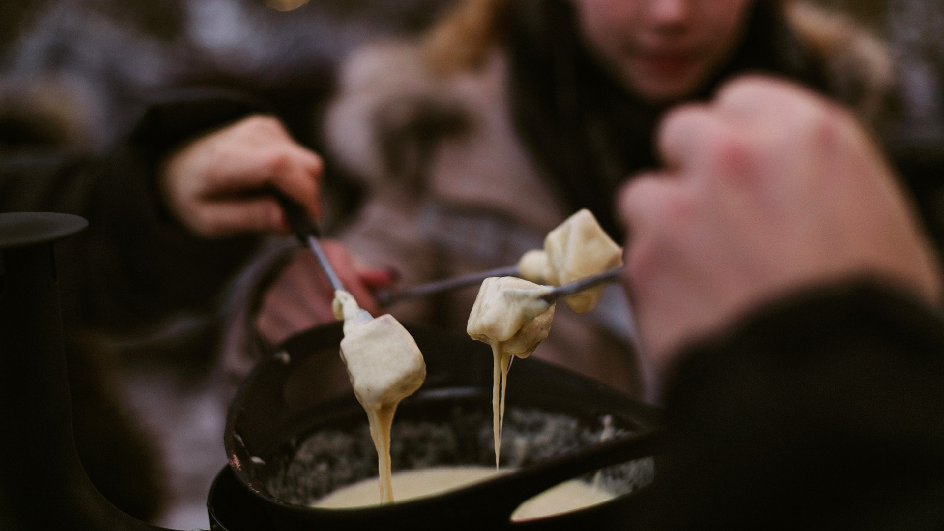 Le fromage fondu extrait du caquelon à fondue. Quatre fourchettes tournent leur morceau de pain dans le caquelon. L’ambiance est hivernale. 