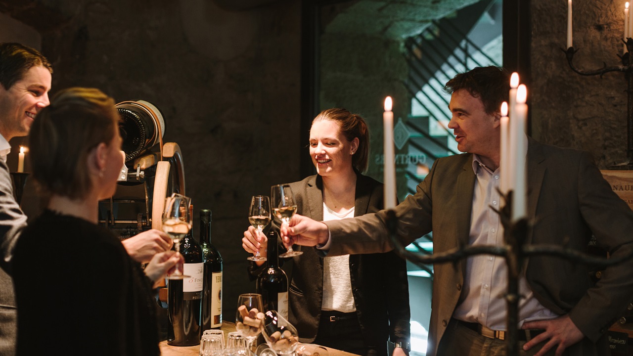 Quatre personnes trinquent dans la cave à vin avec un verre de vin blanc. Une bougie scintille à l’arrière-plan, de nombreuses bouteilles de bons vins sont disposées sur la table. 
