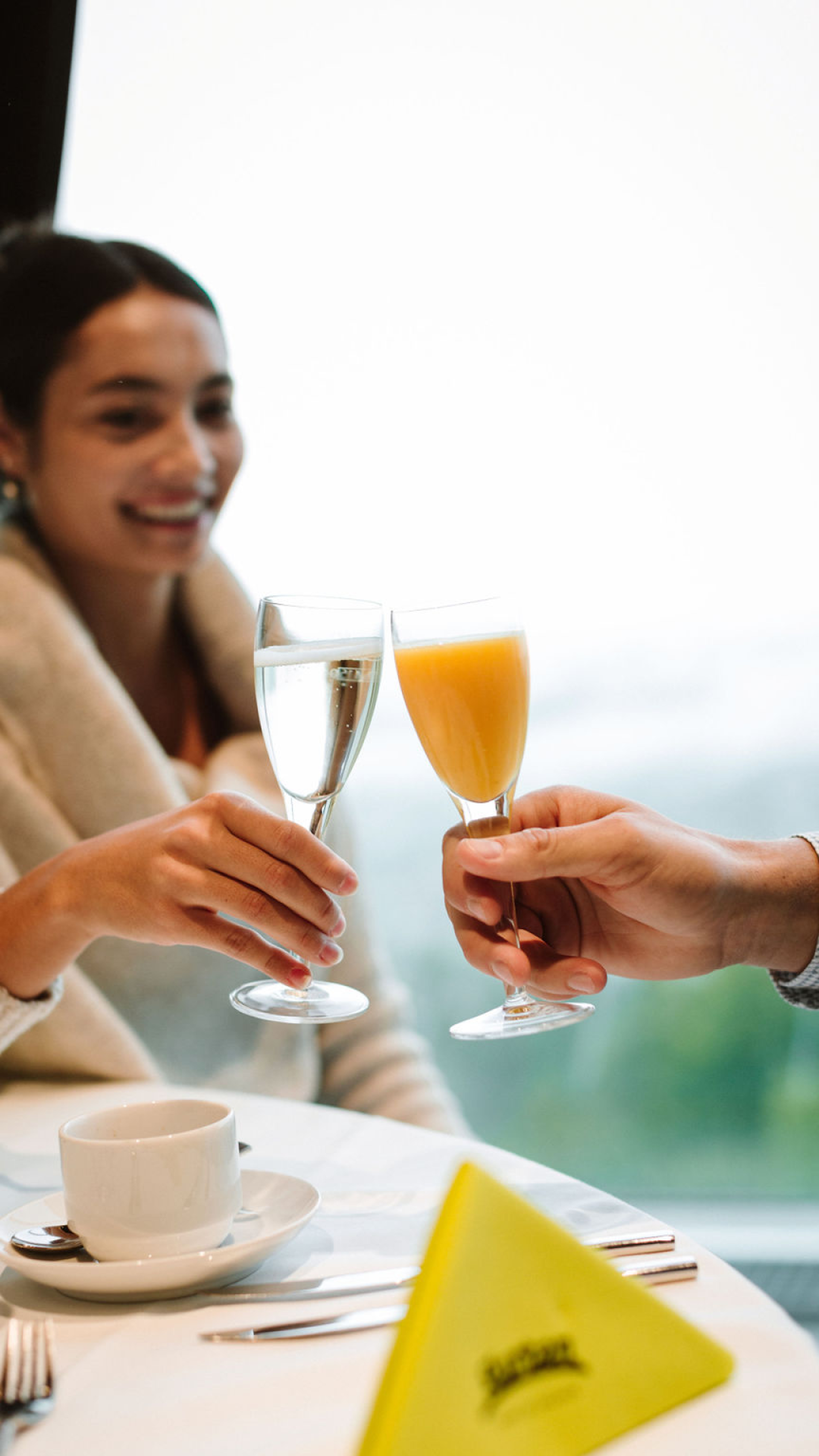 Two people toasting with champagne glasses. One person is toasting with orange juice and the person visible on the left is toasting with champagne.