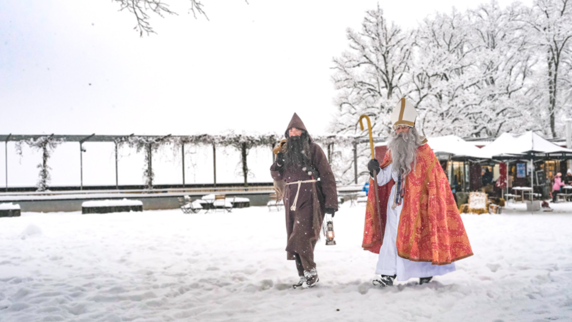Samichlous and Schmutzli walk through the snow-covered meadow with lots of children in the background.