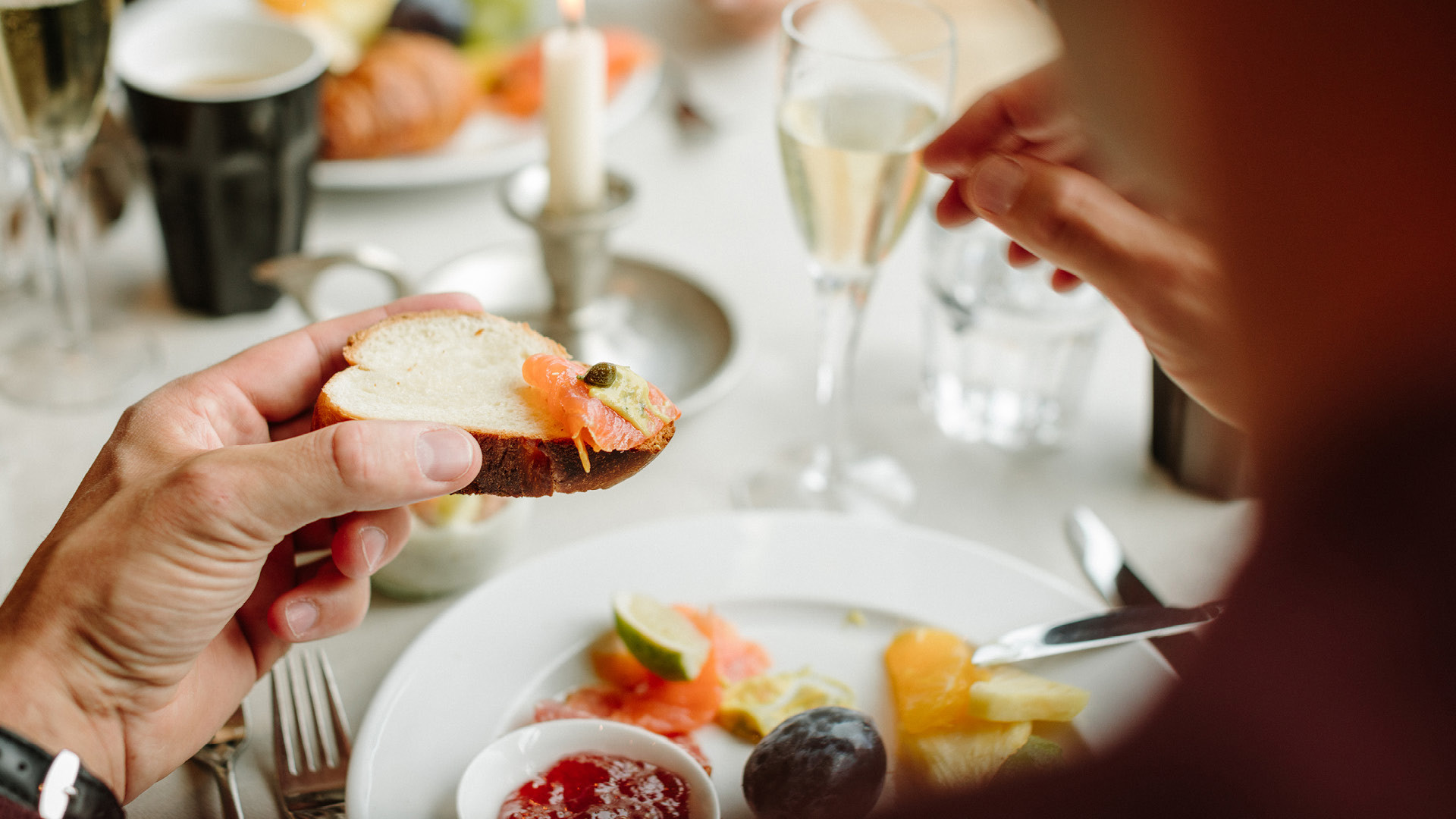 Une personne tient dans sa main un morceau de pain garni de saumon lors d'un Sunntigsbrunch. Son assiette contient encore plus de saumon et des fruits.