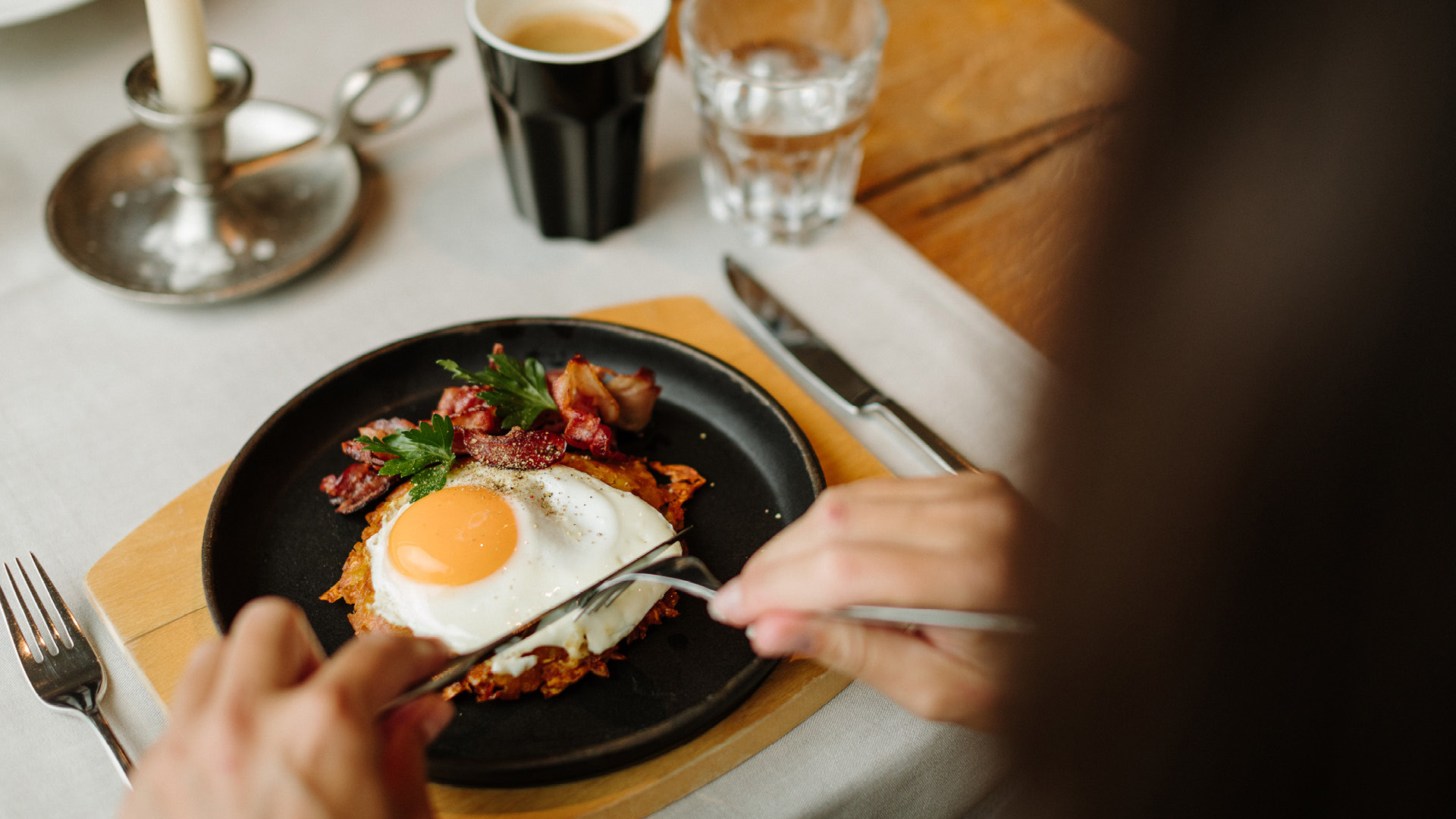 Une personne qui coupe un œuf au plat. Il est posé sur une assiette noire et une nappe blanche.
