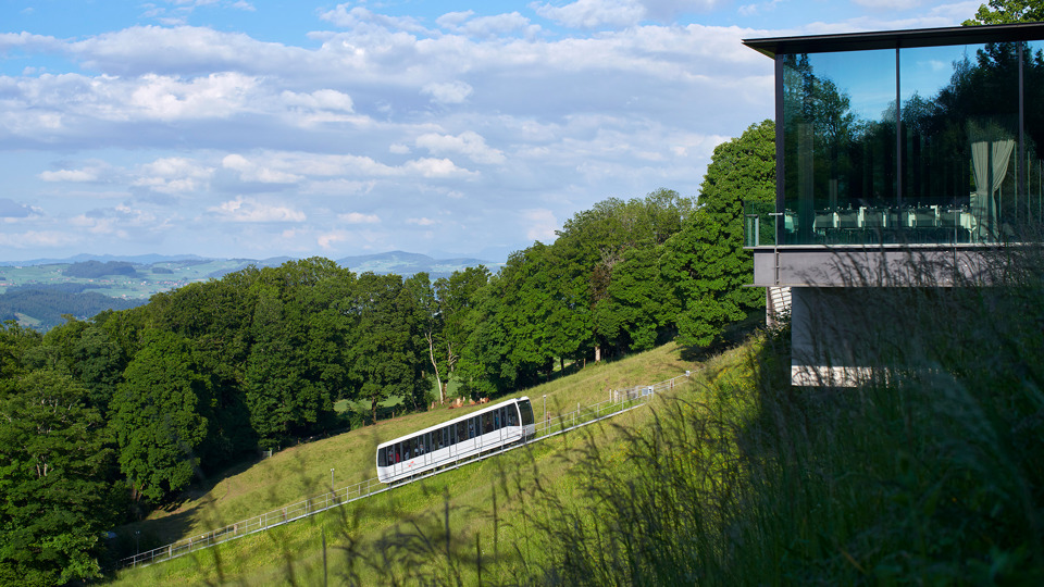 Le funiculaire Gurtenbahn devant une forêt et à côté du Pavillon, un lieu événementiel, photographié depuis l'herbe.