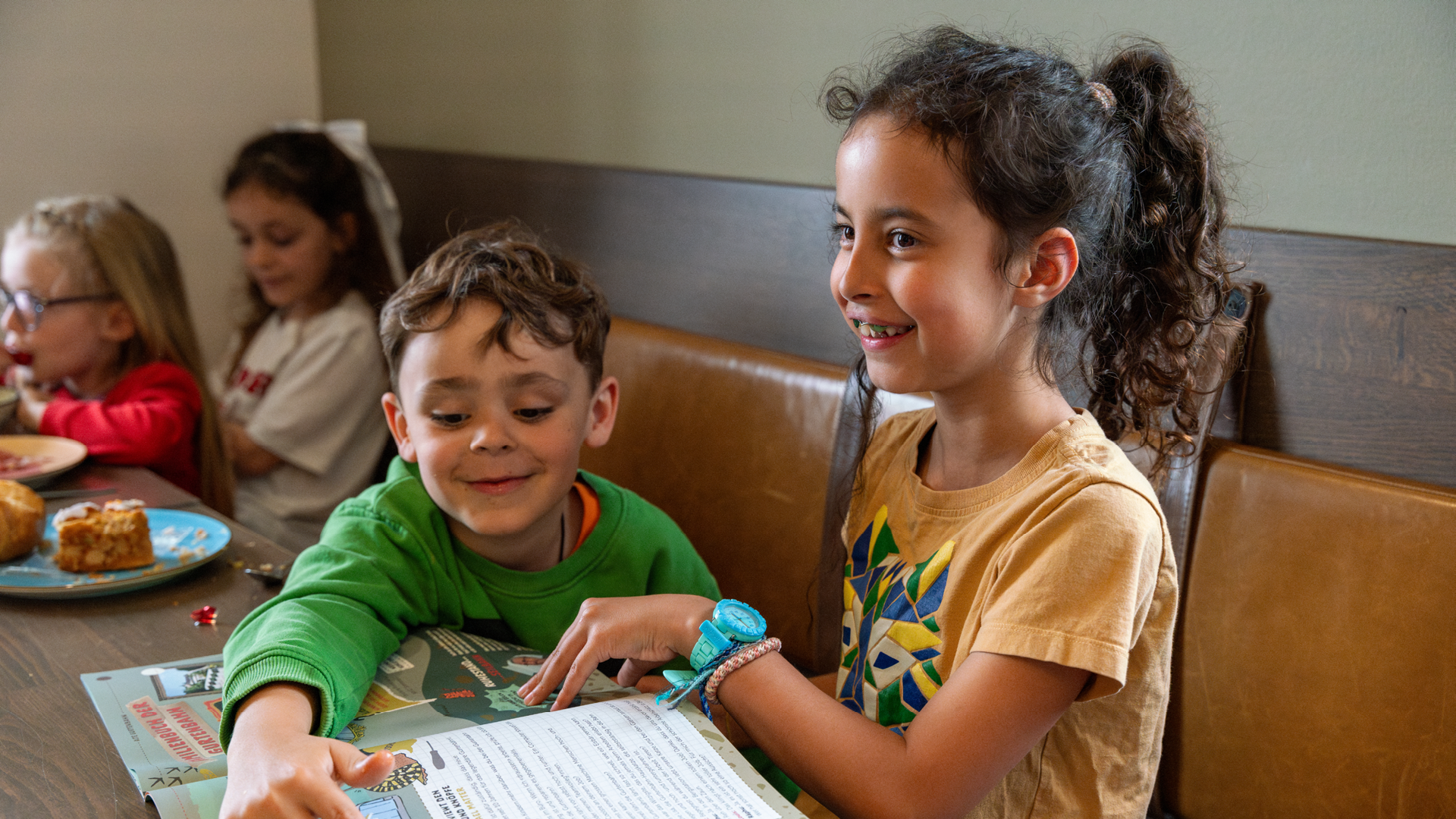 Four children sitting at a table having brunch in the Huusbüffe. The two at the back are eating and the two at the front are enjoying themselves with the Chinderzytig.