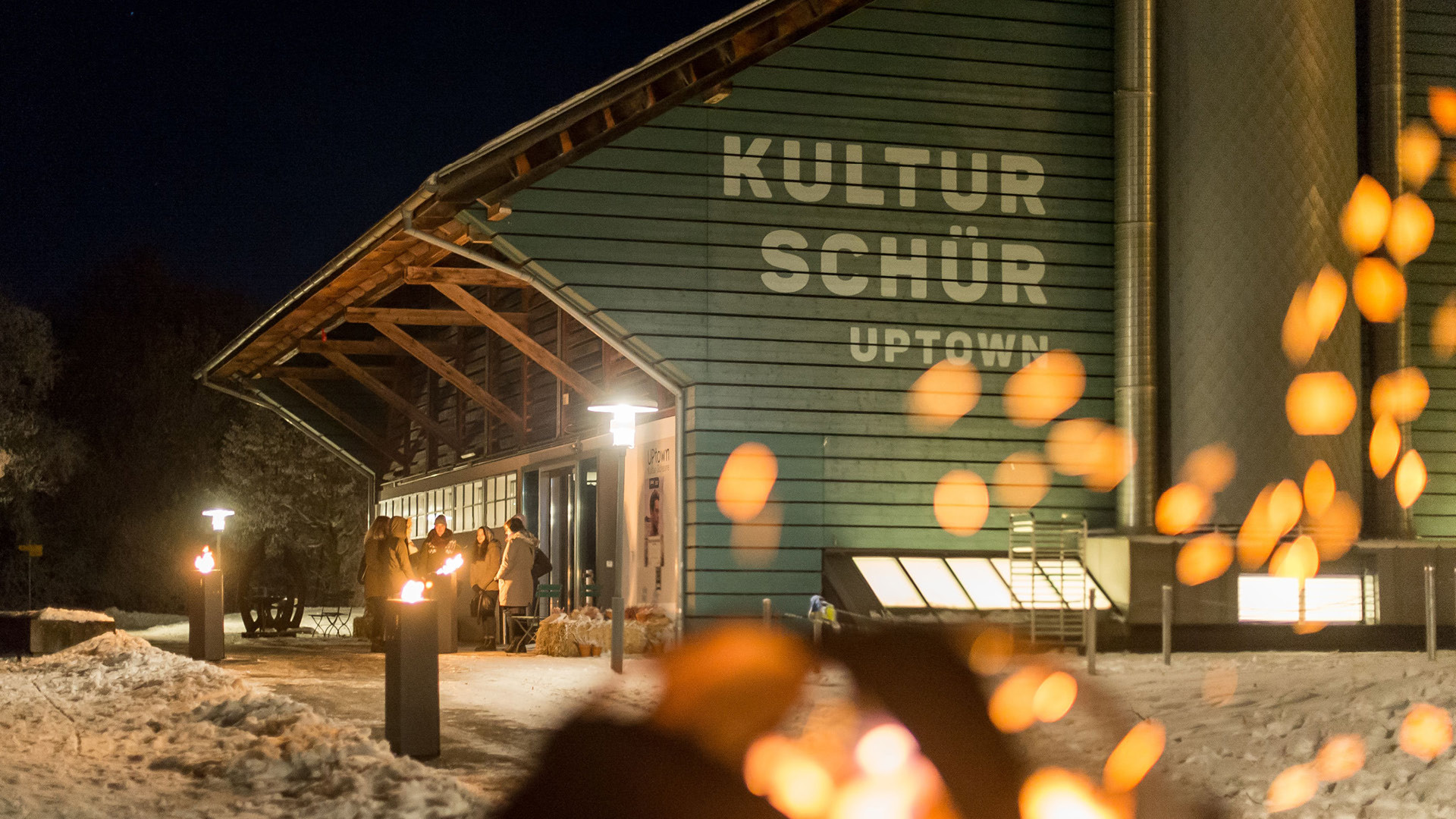 Die Eventlokalität Schüür im Winter mit einer Personengruppe vor dem Eingang und gemütlichen Feuerstellen am Weg entlang.