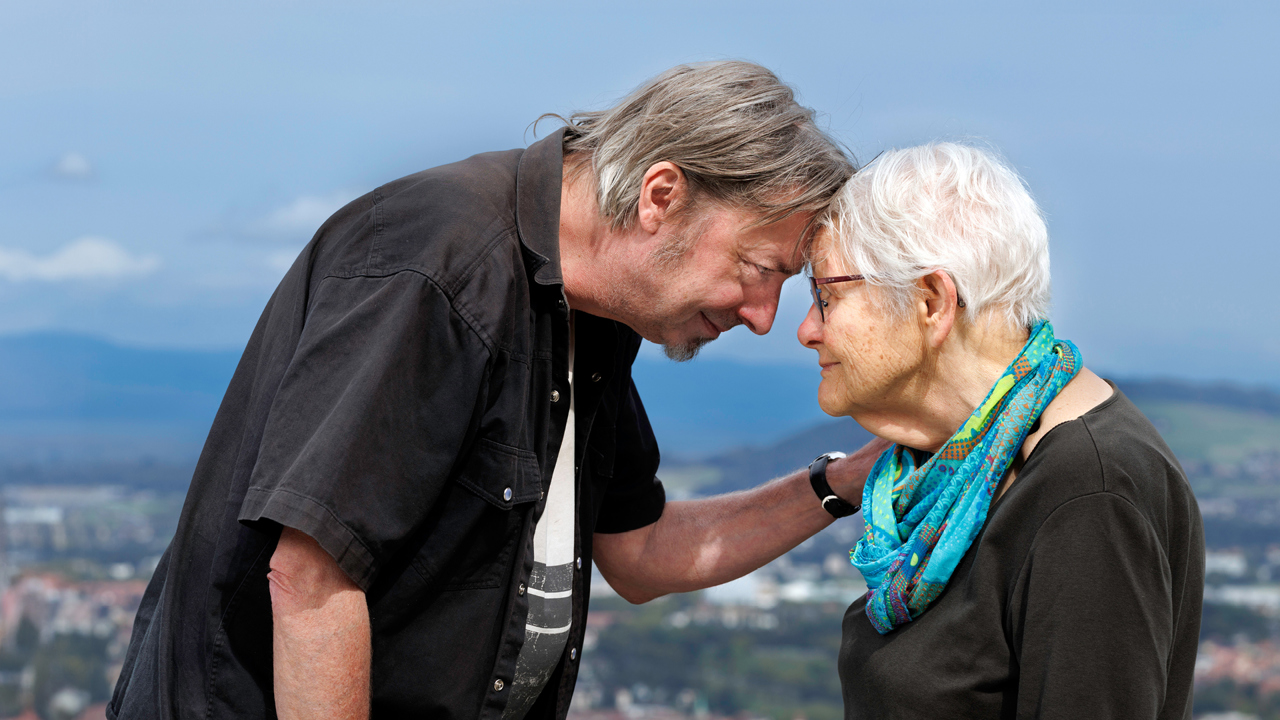 Eine ältere Frau und ihr erwachsener Enkel stehen Stirn an Stirn vor der Aussicht vom Gurten auf Bern.