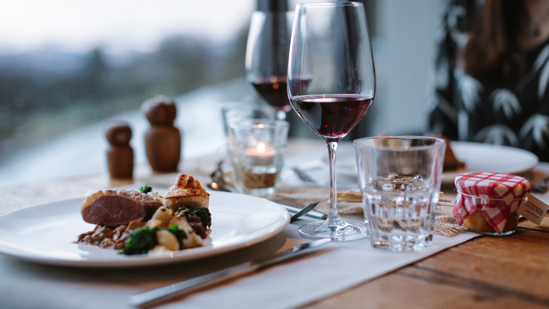 A table at Gurtners restaurant with two plates, two wine glasses, and a welcome gift. The plates are filled with meat and vegetables.