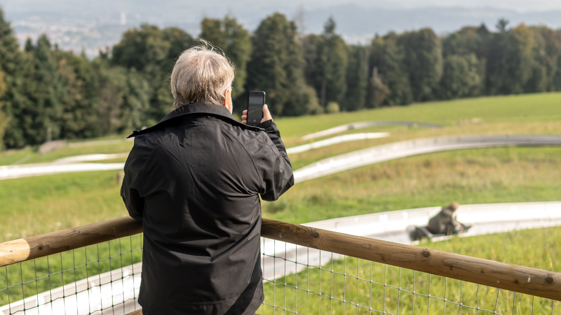 Grandfather from behind, photographing his grandson sledding at the photo point on the toboggan run