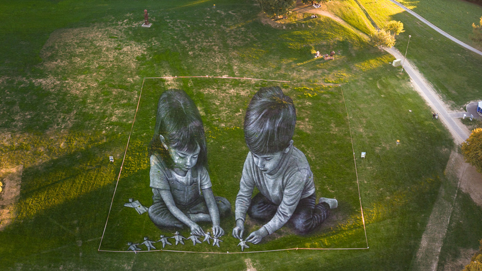 Monumental fresco by Saype depicting two children laying out origami figures.