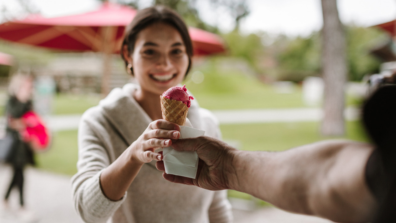 Eine Person welche einer anderer Person ein Eis in die Hand gibt. Die Person trägt einen beigen Pullover.