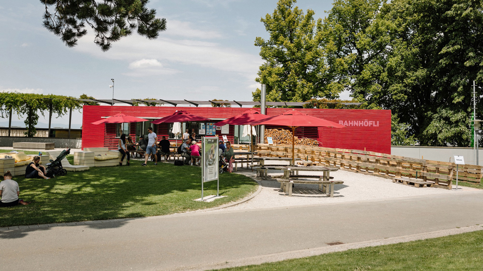 Le restaurant Bahnhöfli. Il est peint en rouge, dispose de pare-soleil rouges près des tables et se trouve à côté d'une pelouse verte.