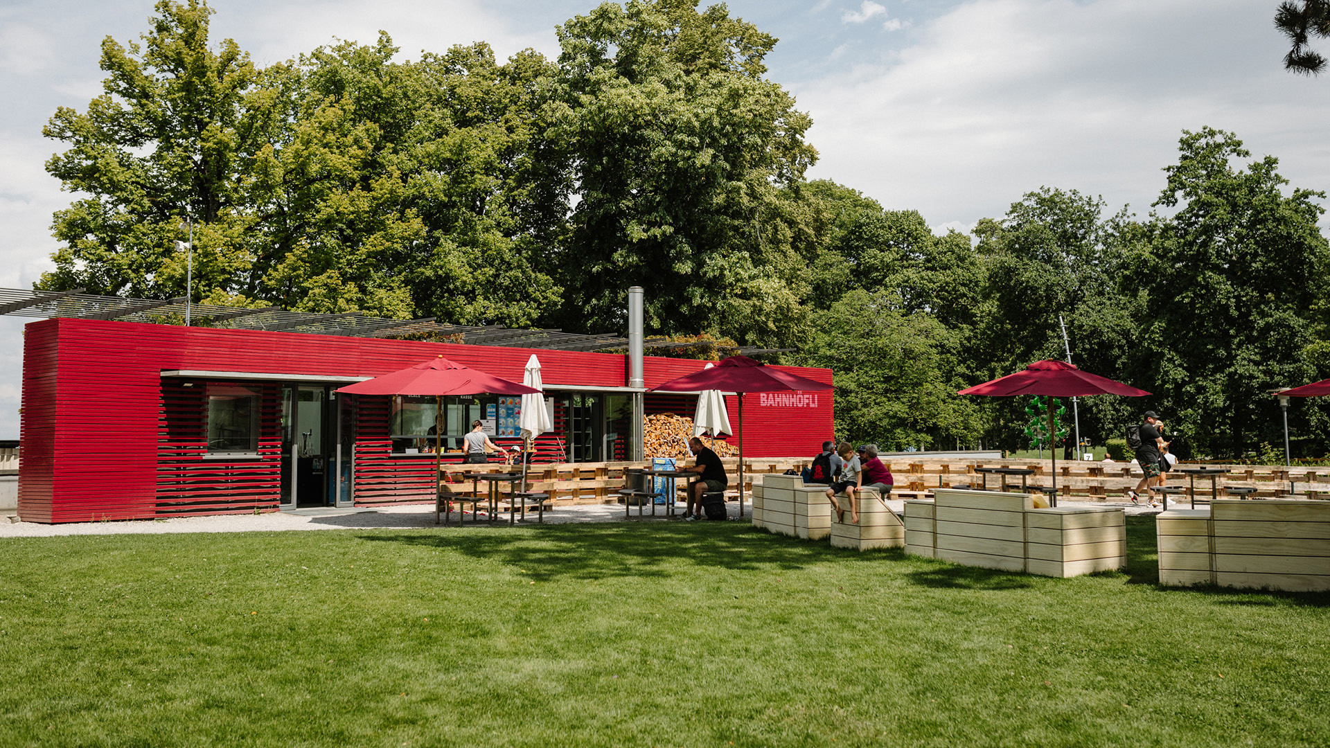 Le restaurant Bahnhöfli. Il est peint en rouge, dispose de pare-soleil rouges près des tables et se trouve à côté d'une pelouse verte.