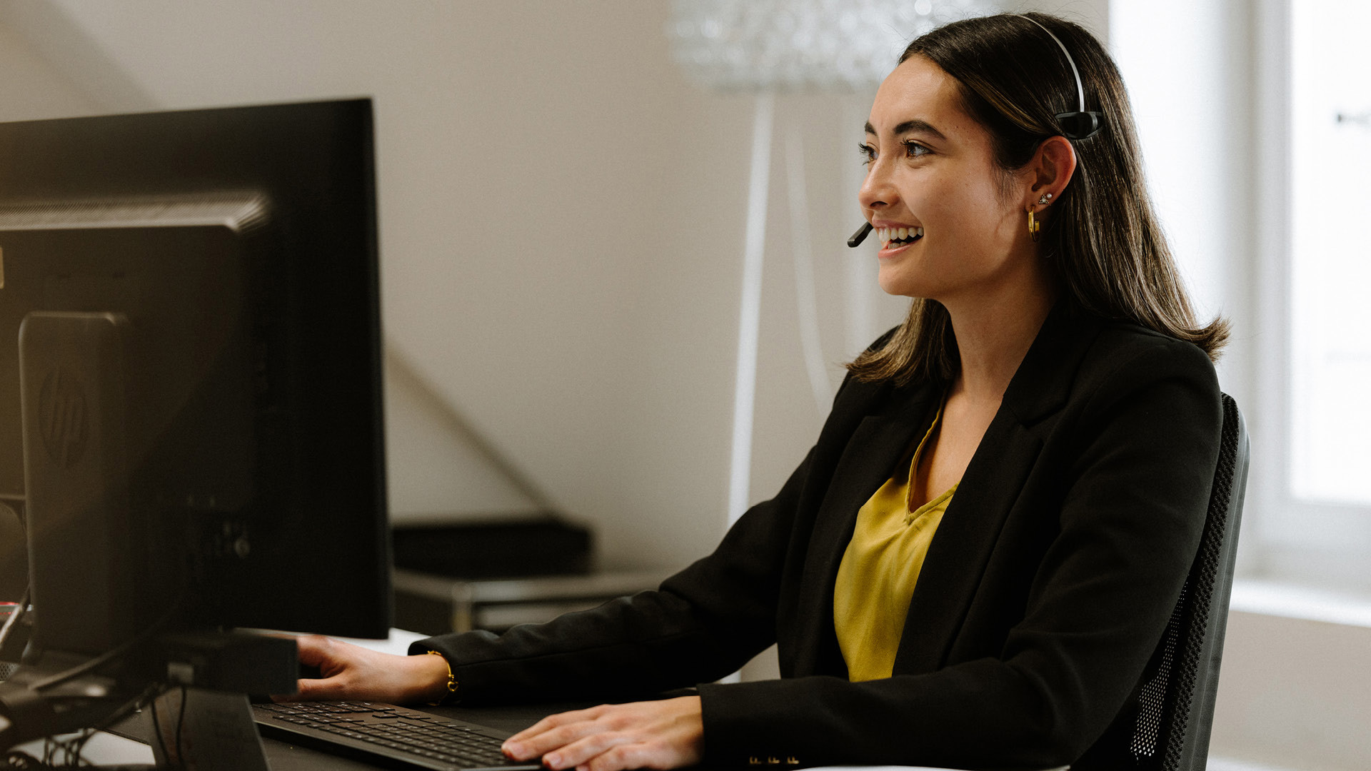 A person wearing a yellow blouse and a black blazer is sitting at a desk with a computer. They are wearing a headset and typing something on the keyboard.