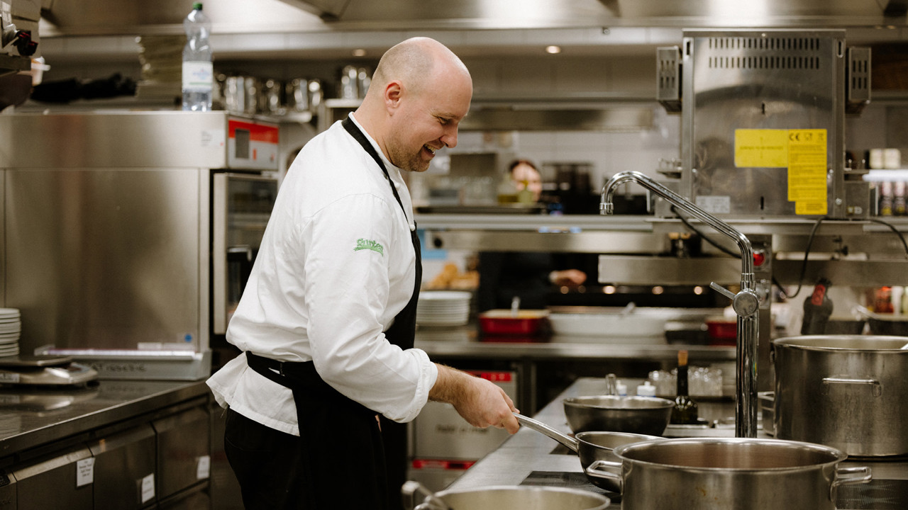 Un cuisinier vêtu d'une veste blanche et d'un tablier noir, qui fait sauter une poêle. Il se tient devant la plaque de cuisson dans la cuisine du Gurtners.