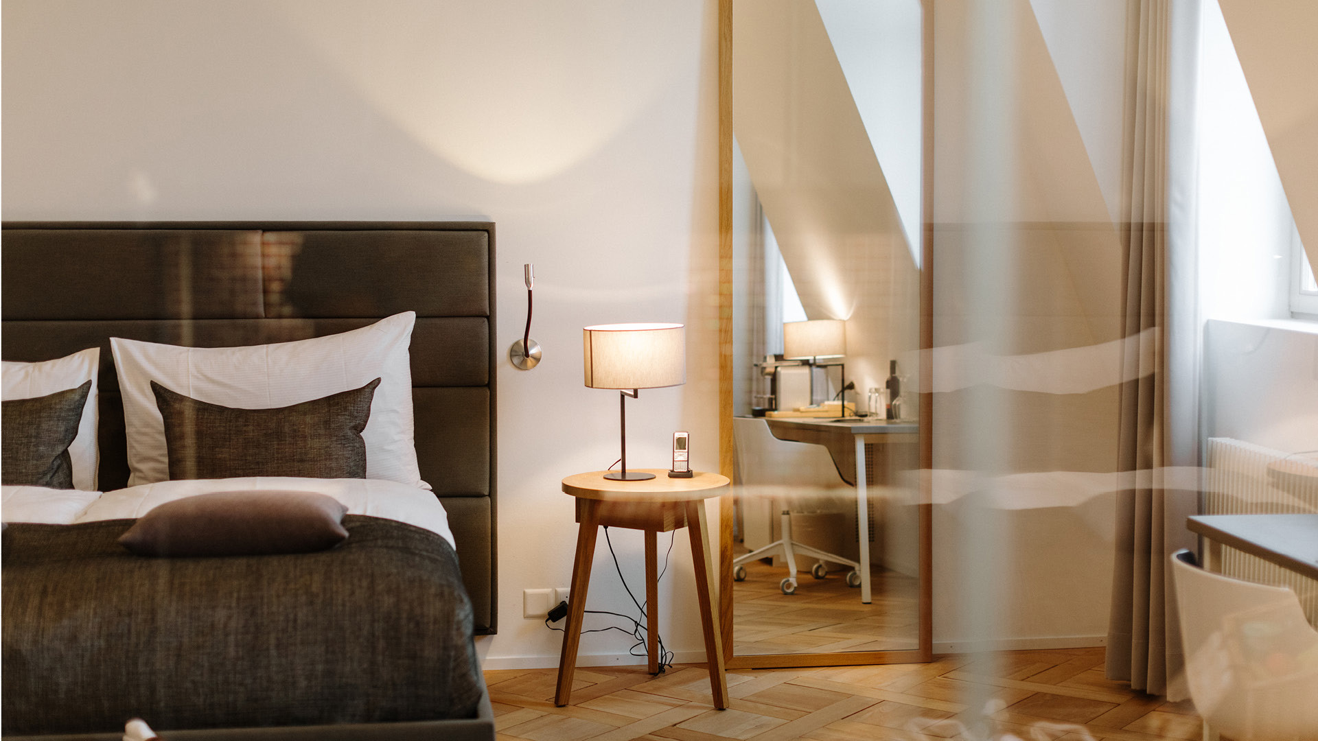 Eine Nahaufnahme eines Hotelzimmers mit einem grauen Bett mit weissem Überzug, einem braunen Nachttisch und einer Lampe und einem Spiegel.