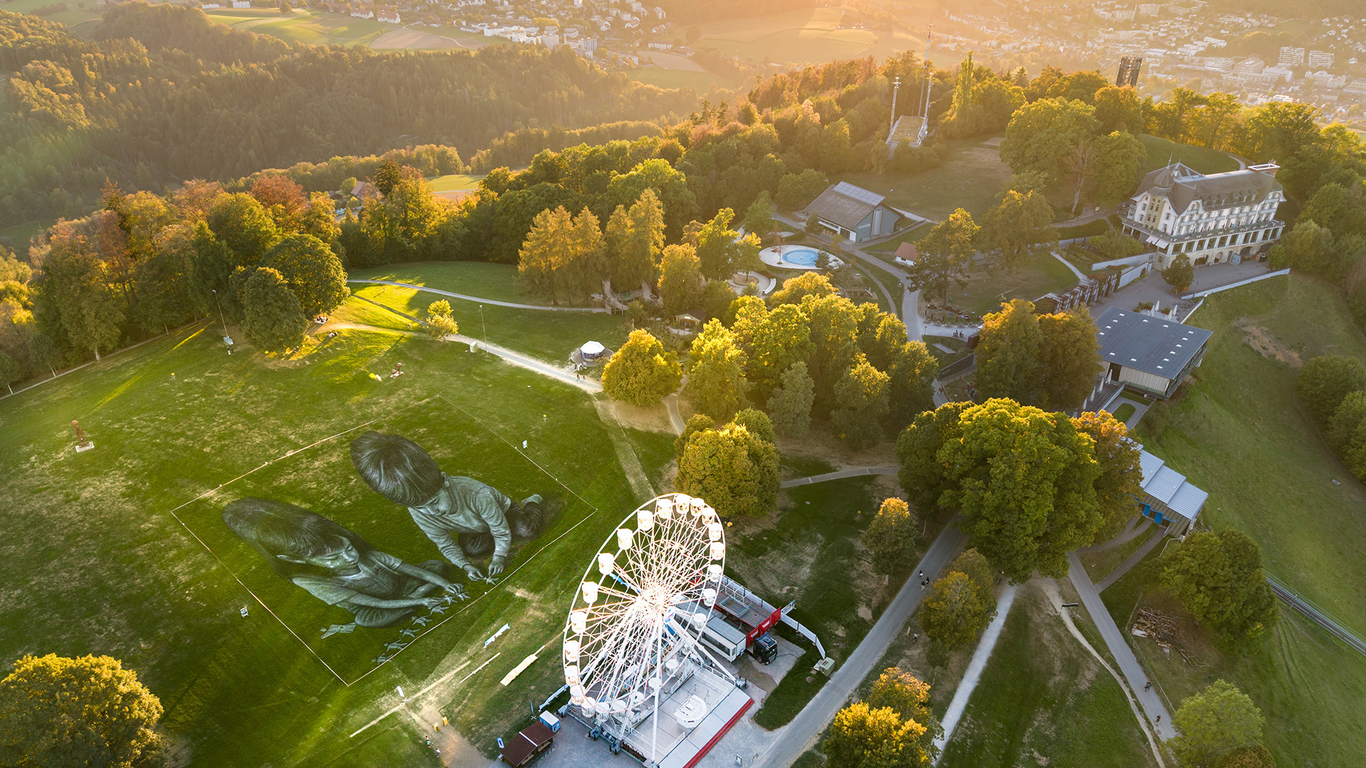 Saype-Kunstwerk und Riesenrad auf dem Gurten im goldenen Abendlicht.