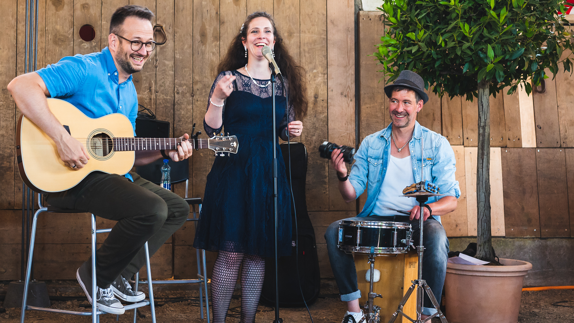 Une chanteuse, un guitariste et un batteur devant un mur en bois