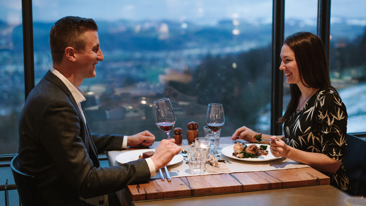 Two people sitting at a table during a candlelight dinner. The person on the right is holding a fork, and the person on the left is lifting a glass.