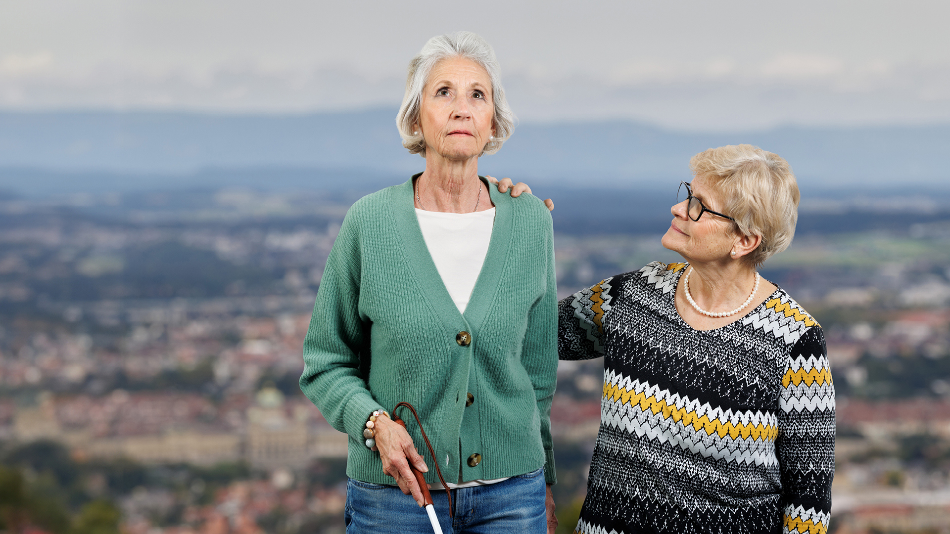 Eine ältere Frau mit einem Blindenstock wird von einer zweiten älteren Frau an der Schulter berührt. Dahinter die Aussicht vom Gurten auf das Bundeshaus.