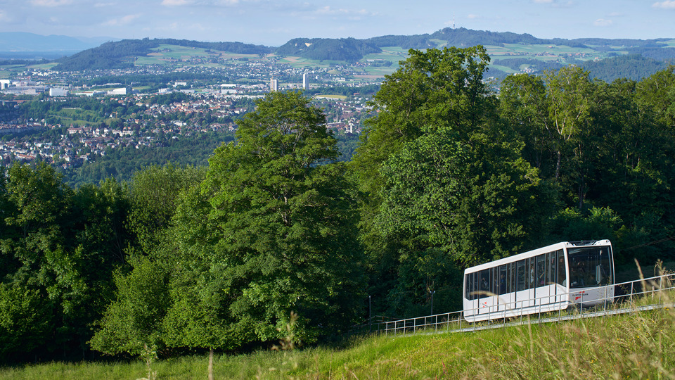 Une photo aérienne du funiculaire Gurtenbahn prise de jour, devant des arbres, avec vue sur la ville de Köniz.