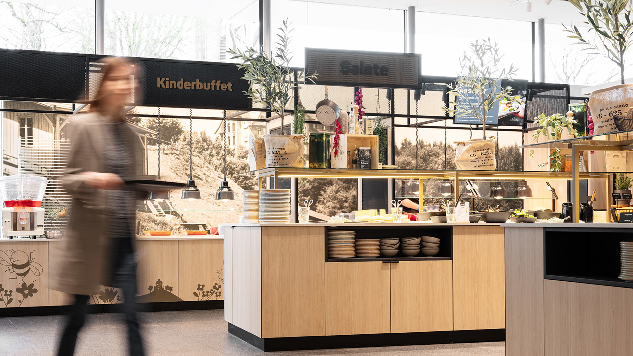 Der Buffetbereich vom Selbstbedienungsrestaurant Huusbüffe mit einer Frau, die ein Serviertablett hält, im Vordergrund.