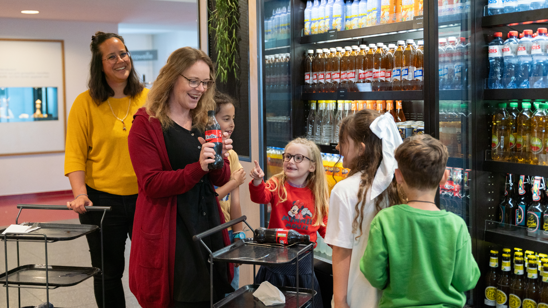Five people taking drinks from the refrigerator shelf. The person wearing a red cardigan is holding a bottle in their hand.