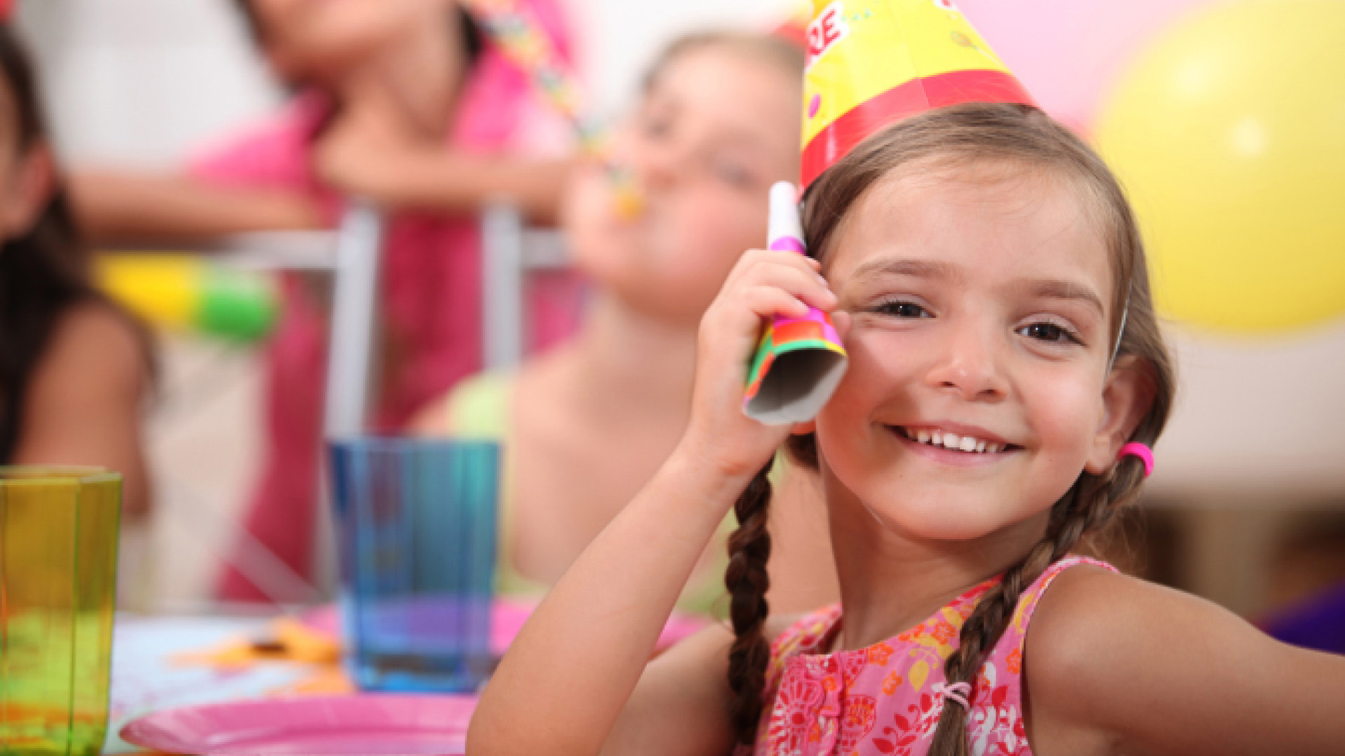 Une petite enfant qui participe à une fête d'anniversaire dans un Huusbüffe. Elle porte une robe rose, un chapeau de fête et deux tresses.