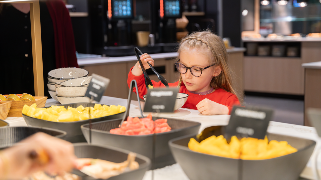 A child with glasses, blonde hair, and a red top, helping themselves to the fruit buffet at brunch in the Huusbüffe.