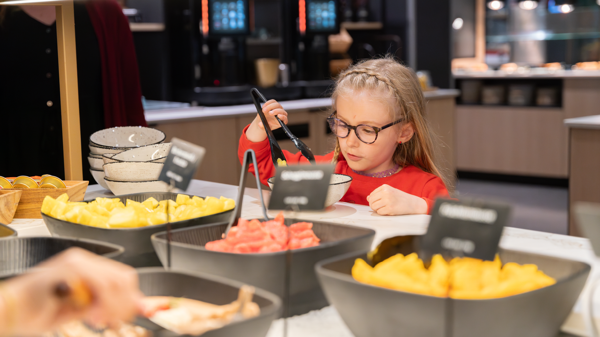 Un enfant portant des lunettes, aux cheveux blonds et vêtu d'un haut rouge, qui se sert au buffet de fruits lors d'un brunch au Huusbüffe.