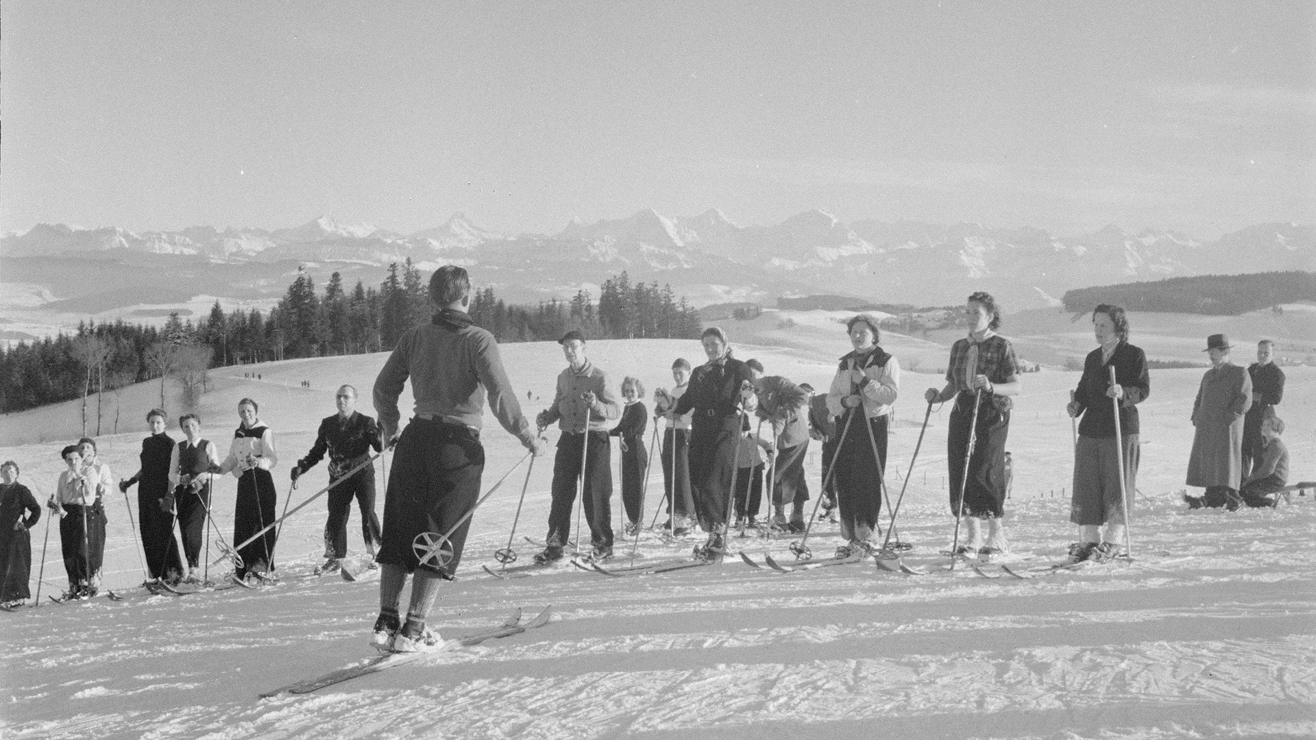 Skifahrer*innen mit alter Skibekleidung und Bergpanorama im Hintergrund auf dem Gurten