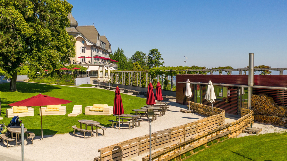 Le restaurant Bahnhöfli. Il est peint en rouge, dispose de pare-soleil rouges près des tables et se trouve à côté d'une pelouse verte.