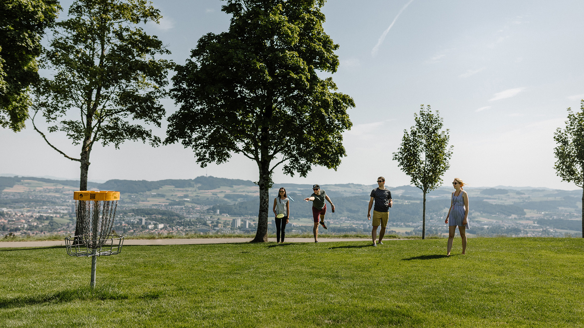Four people are playing disc golf and trying to throw the discs into the basket.